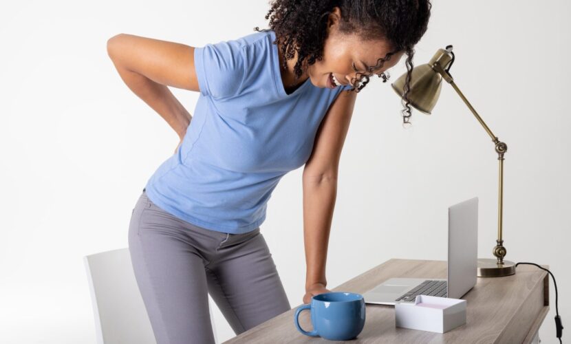 African american mid adult woman with backache standing by laptop, cup and lamp at table, copy space