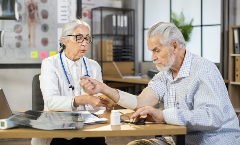 Female middle-aged doctor orthopedist wrapping bandage on arm of senior male patient