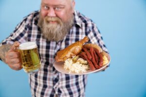 Obese man on blue background, focus on hands with plate of greasy food and beer