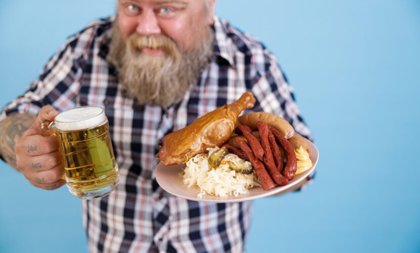 Obese man on blue background, focus on hands with plate of greasy food and beer